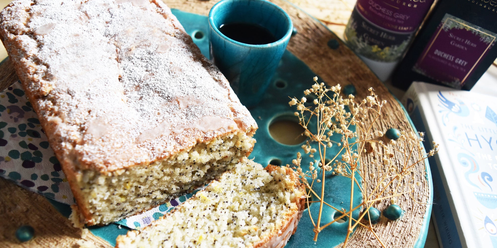 Lemon and Poppy Seed Cake on a plate next to a cup of tea and a PekoeTea Edinburgh Duchess Grey Tea Caddy and PekoeTea Edinburgh Duchess Grey Loose Leaf Box with book about Hygge